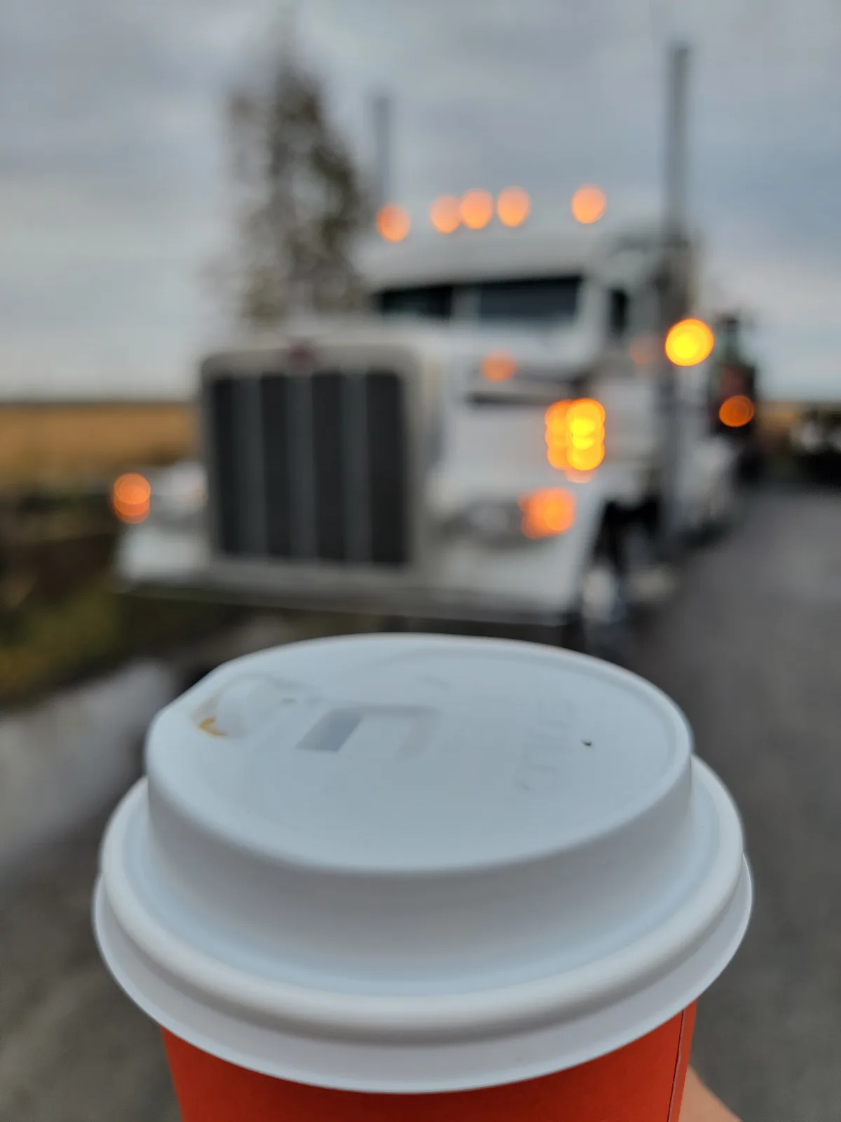 A close-up of a red coffee cup with a white lid is in sharp focus in the foreground, held by a hand. In the blurred…