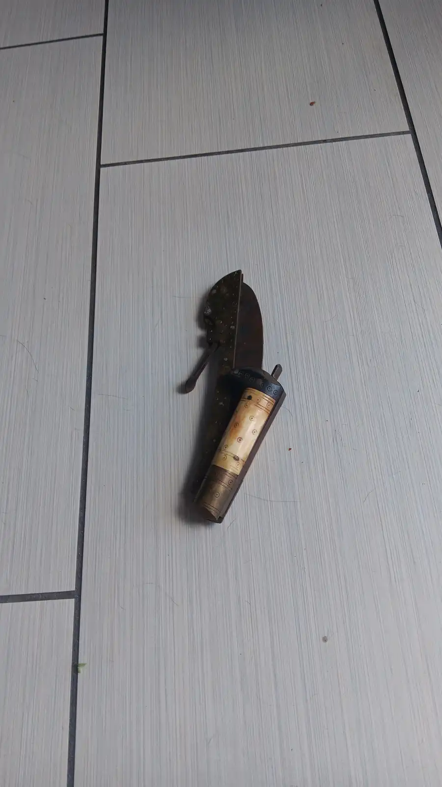 a vintage-style knife with a decorative bone or ivory handle and a metal sheath lies on a light gray tiled floor. The…