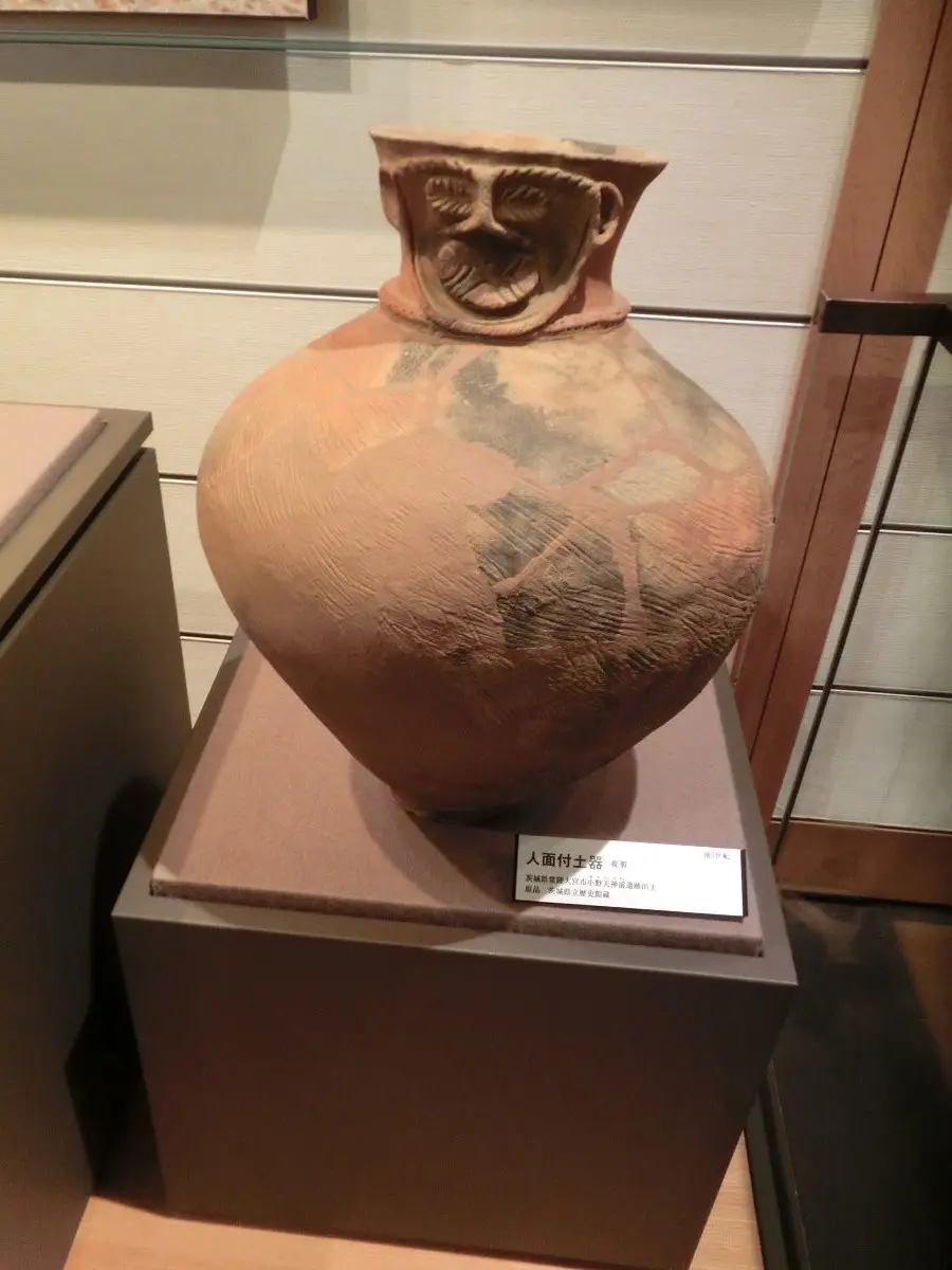 A large, ancient terracotta jar with a human face molded into its neck, displayed on a museum pedestal with a label reading…