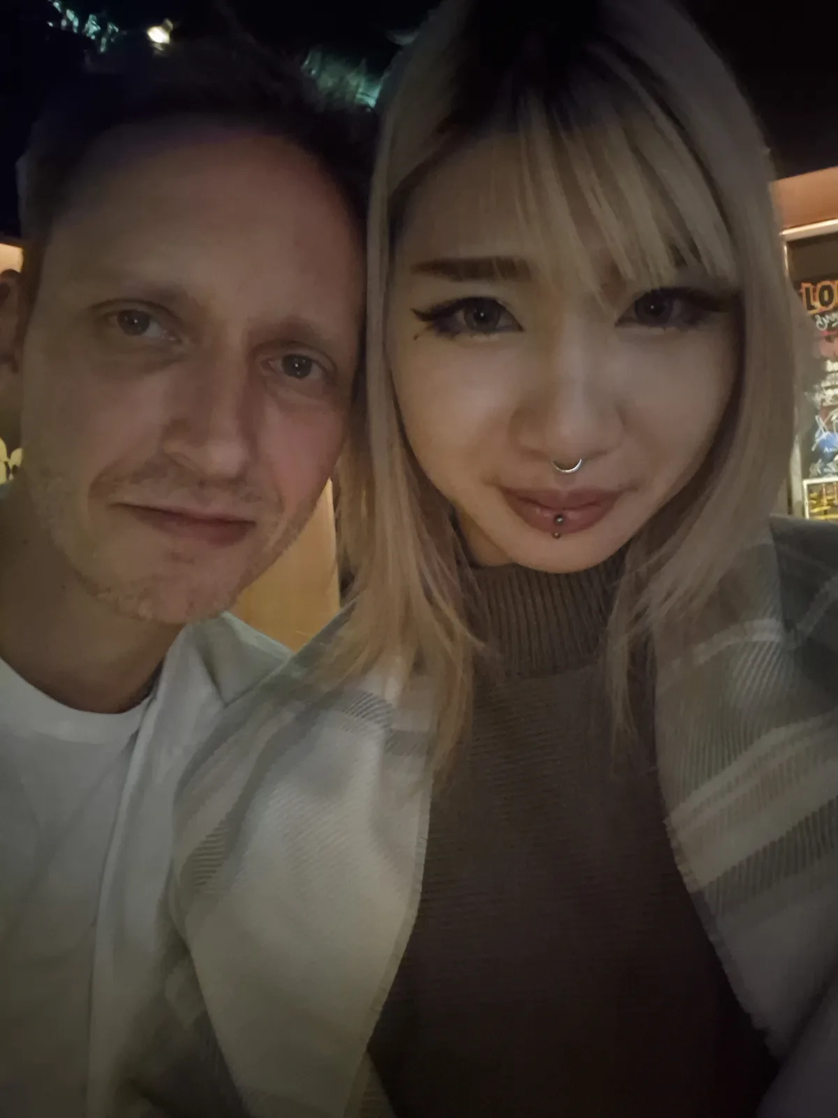 A selfie of a man and a woman at night, the woman has blonde hair, a nose ring, and lip piercing, and is smiling while the…