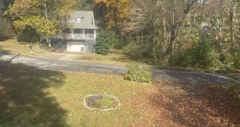 A photo of a suburban house surrounded by autumn trees and fallen leaves, with a small garden bed in the foreground. The…