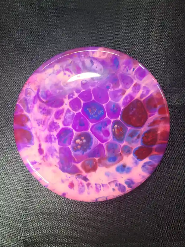A top-down photo of a round, glossy disc with a marbled pattern of purple, pink, and blue colors resembling cellular…