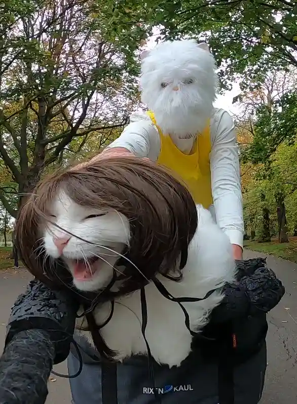A person wearing a fluffy white cat mask and a yellow vest is holding a white cat wearing a brown wig, which has its mouth…