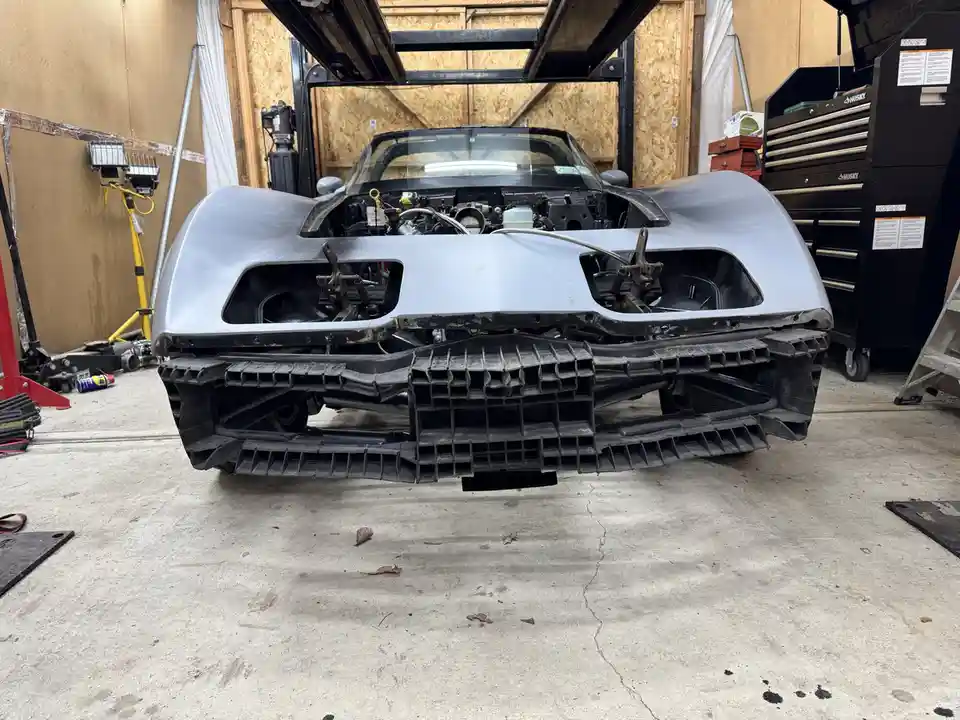 A silver sports car is shown from the front in a garage, with its hood open and engine exposed. The front bumper has been…