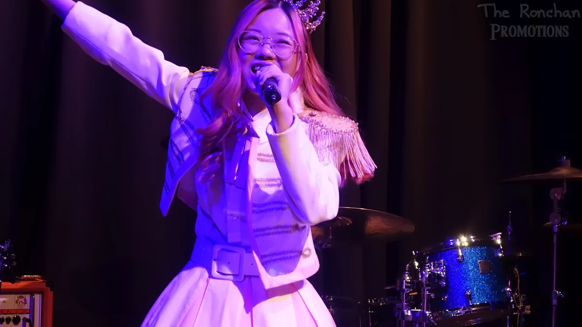 A woman with glasses and a sparkly crown performs on stage in a white fringed outfit, holding a microphone and raising one…