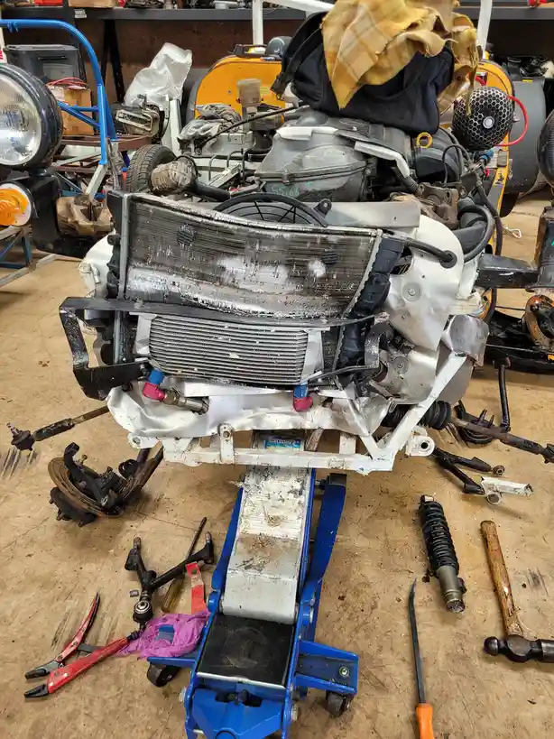 A heavily disassembled car engine and chassis sits on a blue hydraulic jack in a cluttered garage, surrounded by scattered…