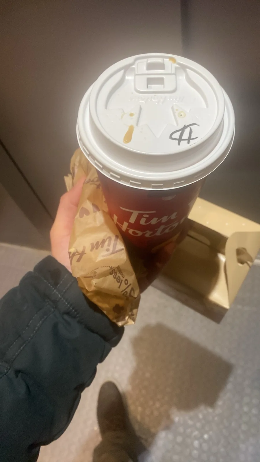 A hand holds a Tim Hortons coffee cup with a white lid that has a black scribble and coffee stains, alongside a brown paper…