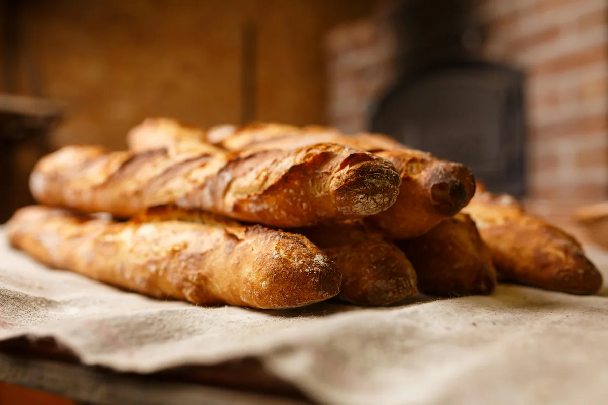 A close-up photograph shows several freshly baked baguettes with golden-brown crusts resting on a textured cloth. The…