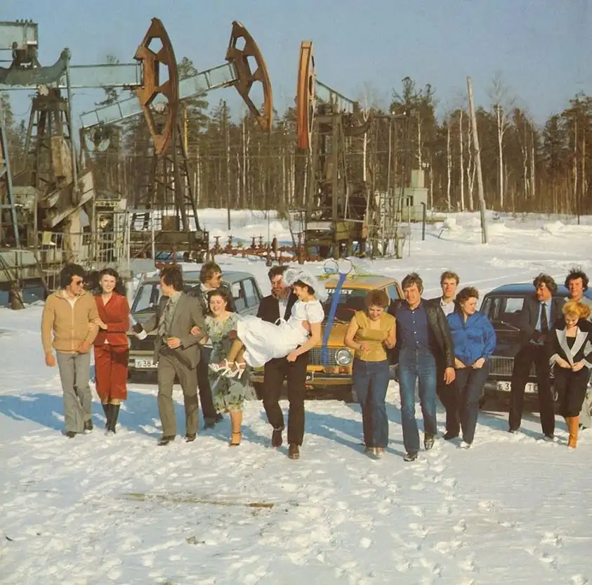 A vintage photo shows a group of people in winter clothing standing in snow near oil pumps and old cars. A man in a suit…