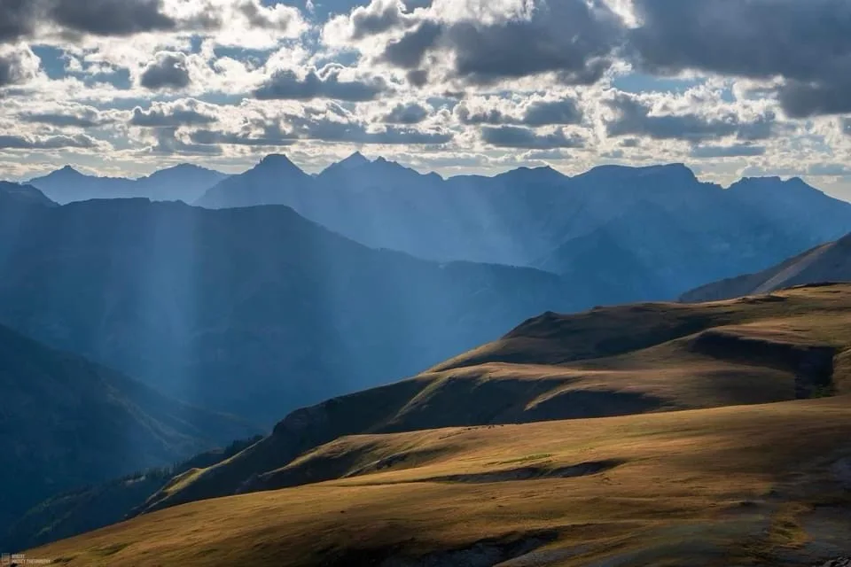 A breathtaking mountain landscape with layered peaks under a cloudy sky, sunbeams piercing through, and golden grassy slopes…
