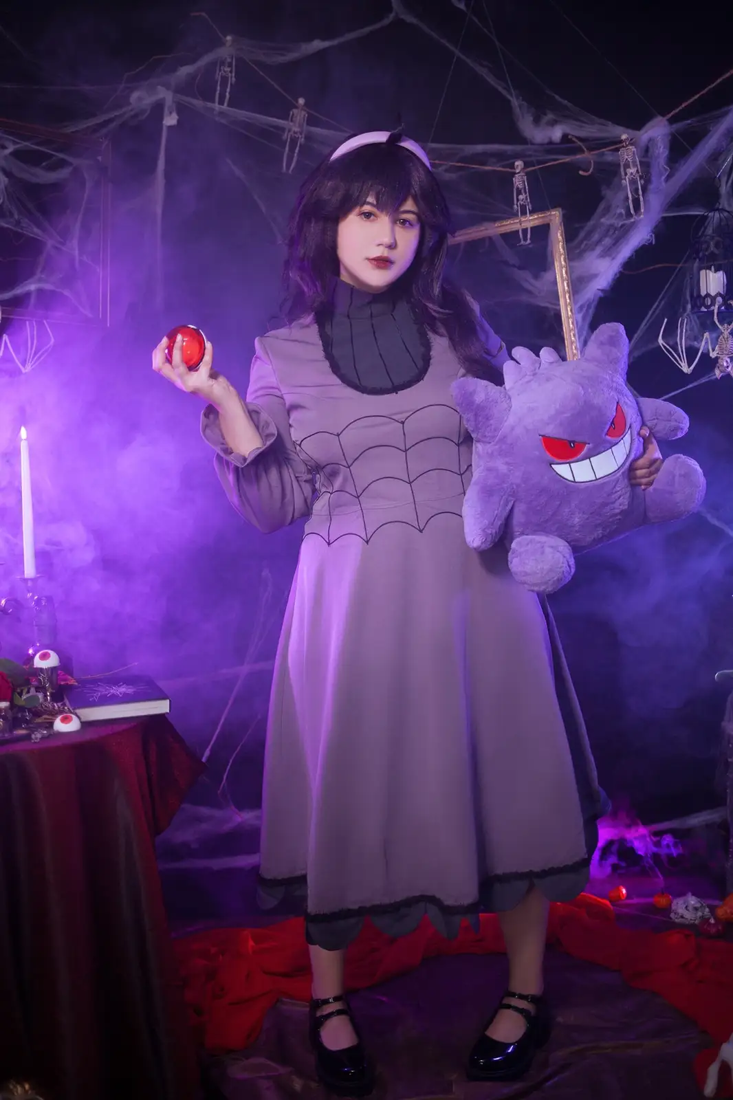 A person in a purple spider-themed dress stands in a dark, spooky room decorated with cobwebs, hanging skeletons, and purple…