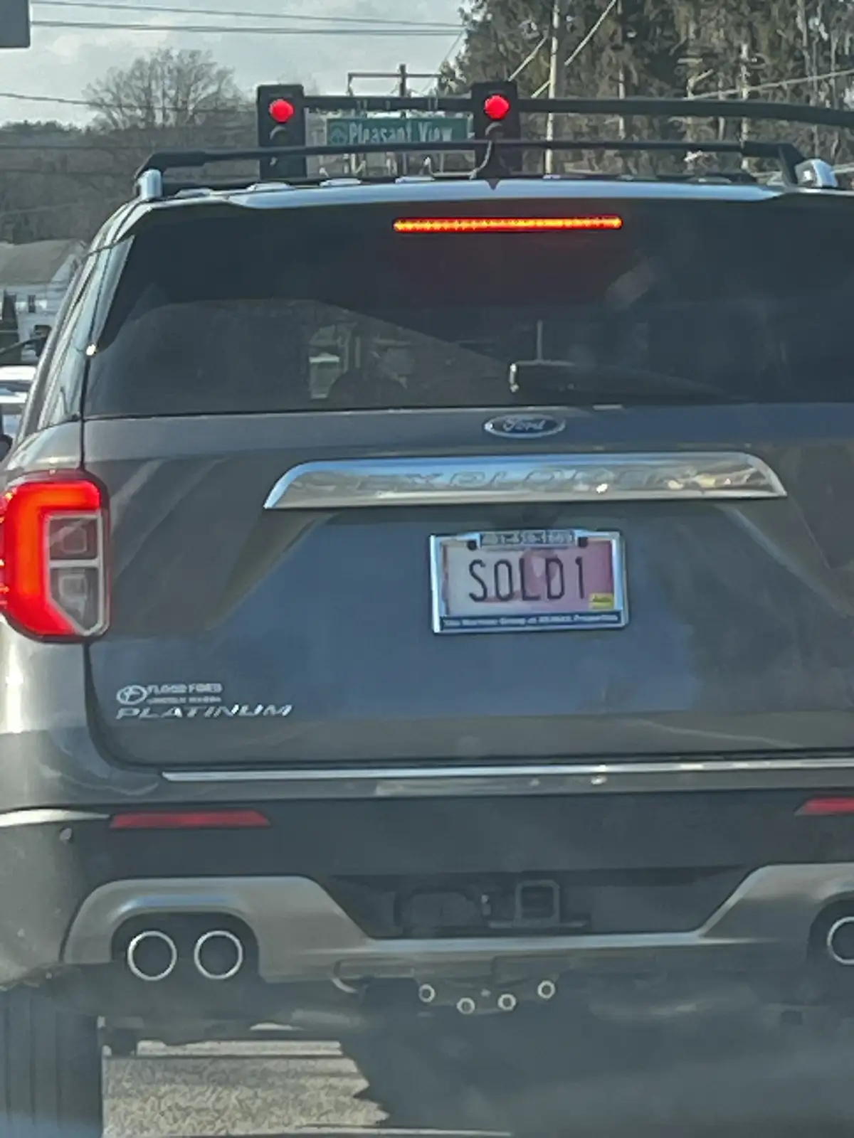 The rear view of a dark gray Ford Explorer Platinum SUV stopped at a red light, showing a custom license plate that reads…