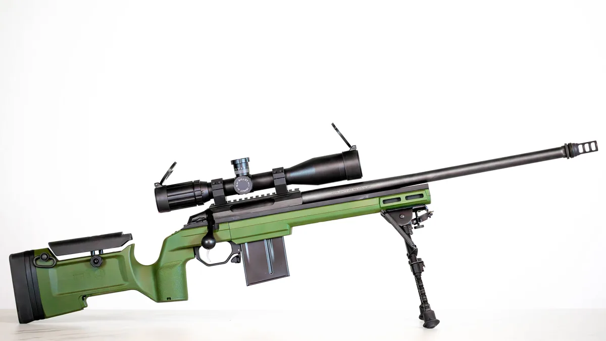 A green and black bolt-action sniper rifle with a large scope and bipod is displayed against a plain white background. The…