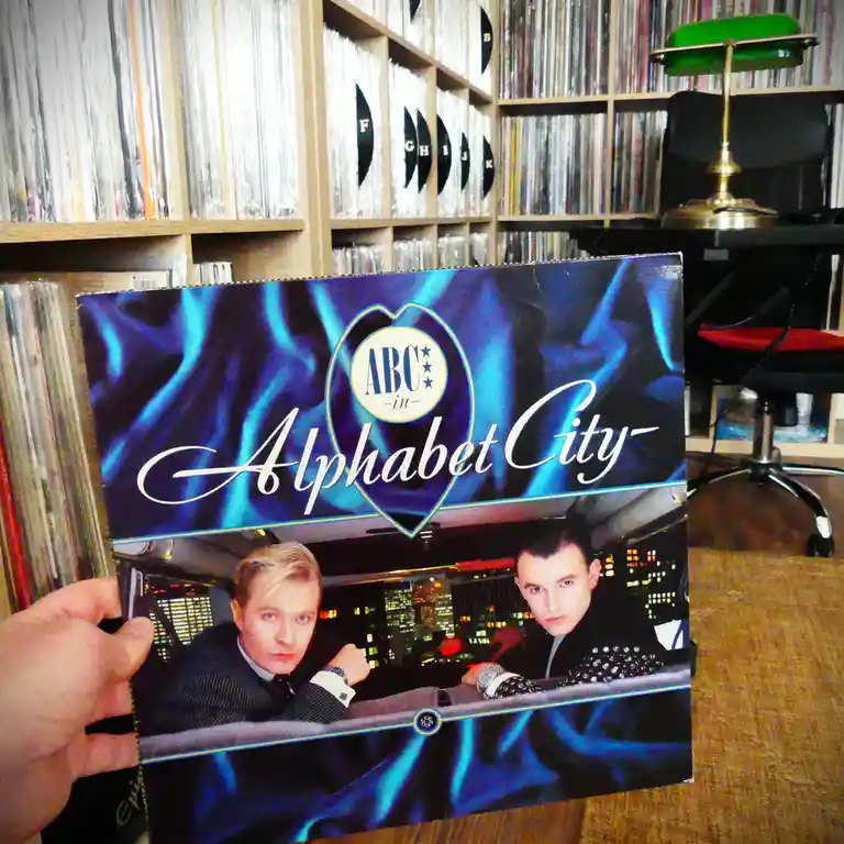 A hand holds up a vinyl record album cover for 'ABC in Alphabet City' featuring two men in suits inside a car with a city…