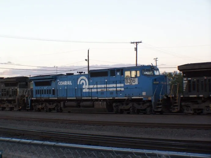 A blue Conrail locomotive, number 8316, sits on train tracks at dusk with power lines overhead and a chain-link fence in the…