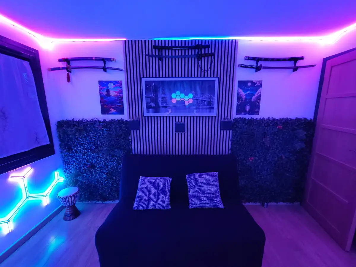 A room with purple and blue neon lighting, featuring a black couch with two patterned pillows, walls adorned with Japanese…