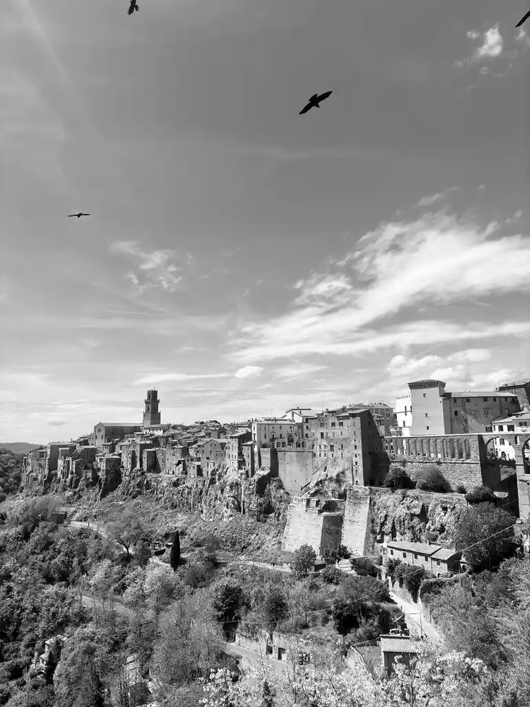 A black-and-white photograph shows a historic European hillside town with stone buildings, a prominent tower, and a bridge…