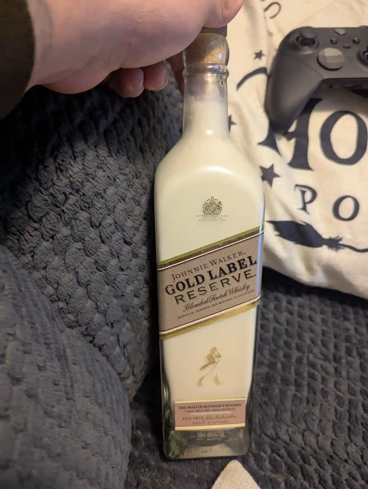 A hand holds a Johnnie Walker Gold Label Reserve whisky bottle that has been filled with a white liquid, likely milk, making…