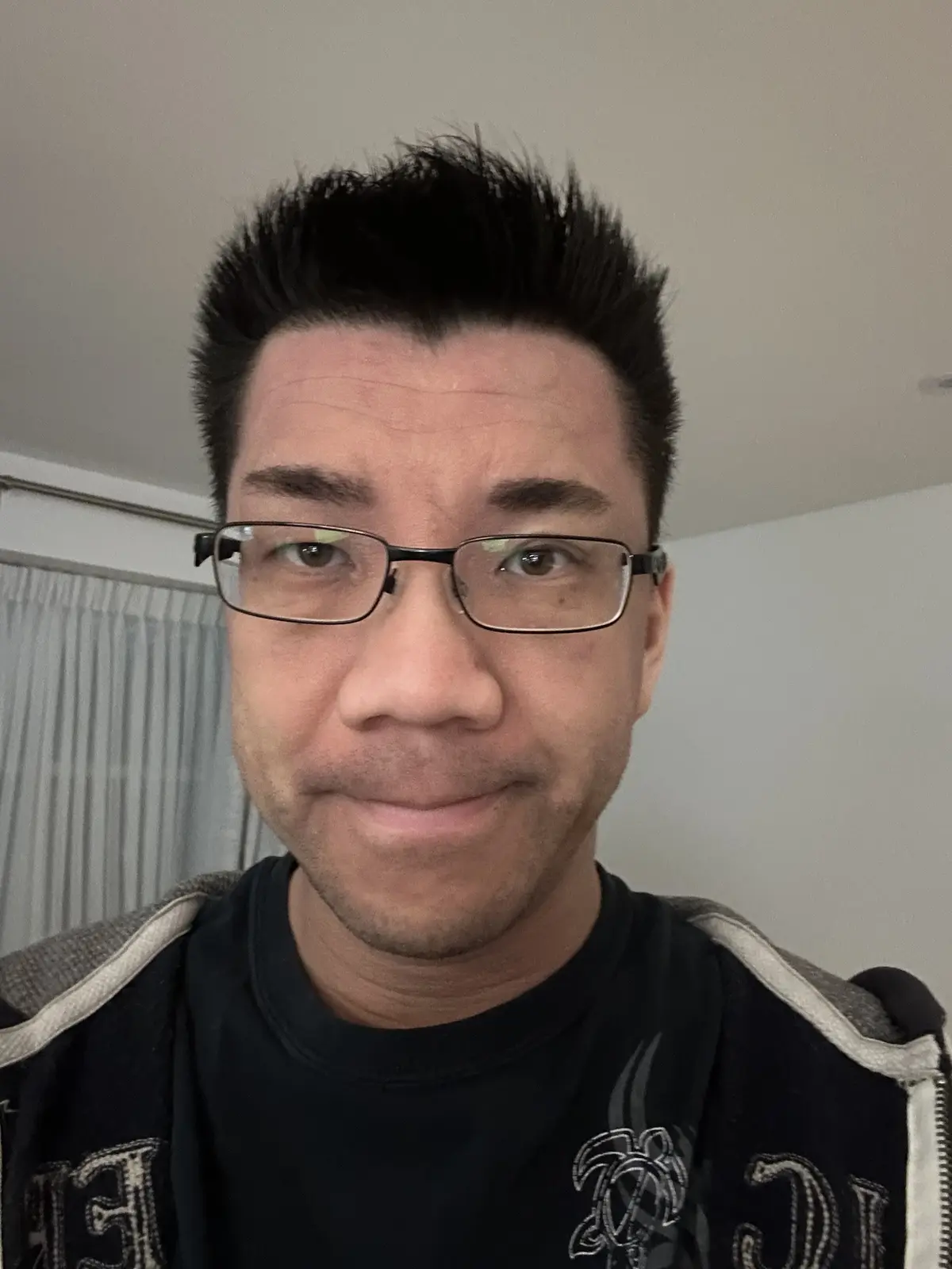 A close-up selfie of a man with dark, spiky hair and glasses, wearing a black shirt under a jacket with grey and white trim…