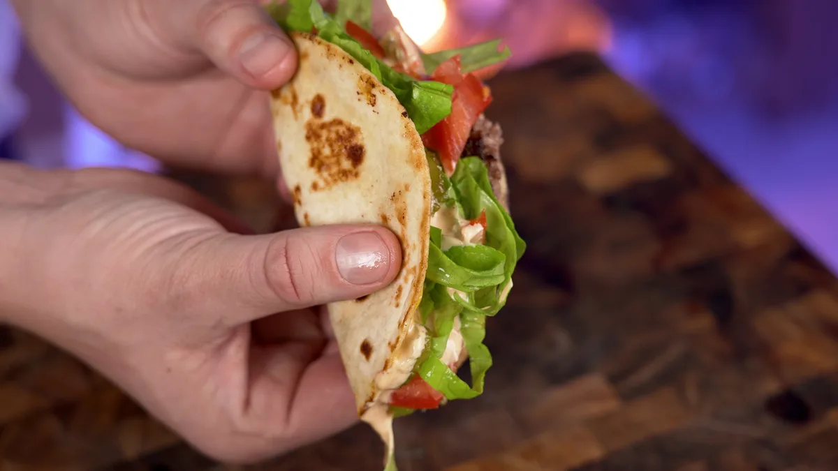 A close-up shot shows two hands holding a taco filled with lettuce, tomato, and a creamy sauce, with the sauce visibly…