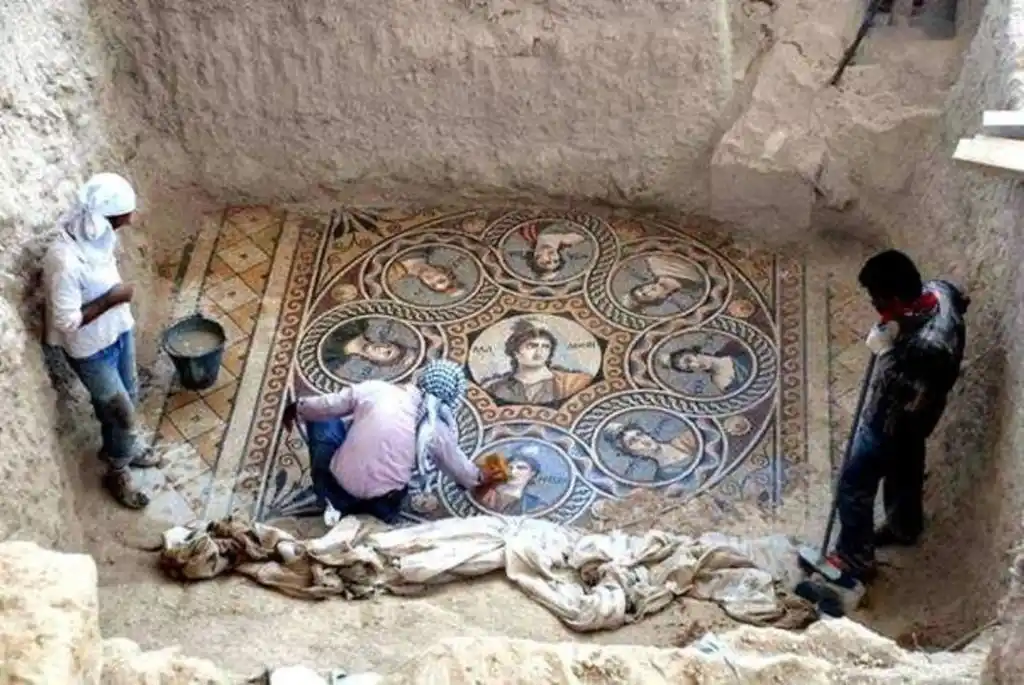 Three archaeologists are excavating an ancient mosaic floor in a dug-out chamber. The mosaic depicts seven circular…