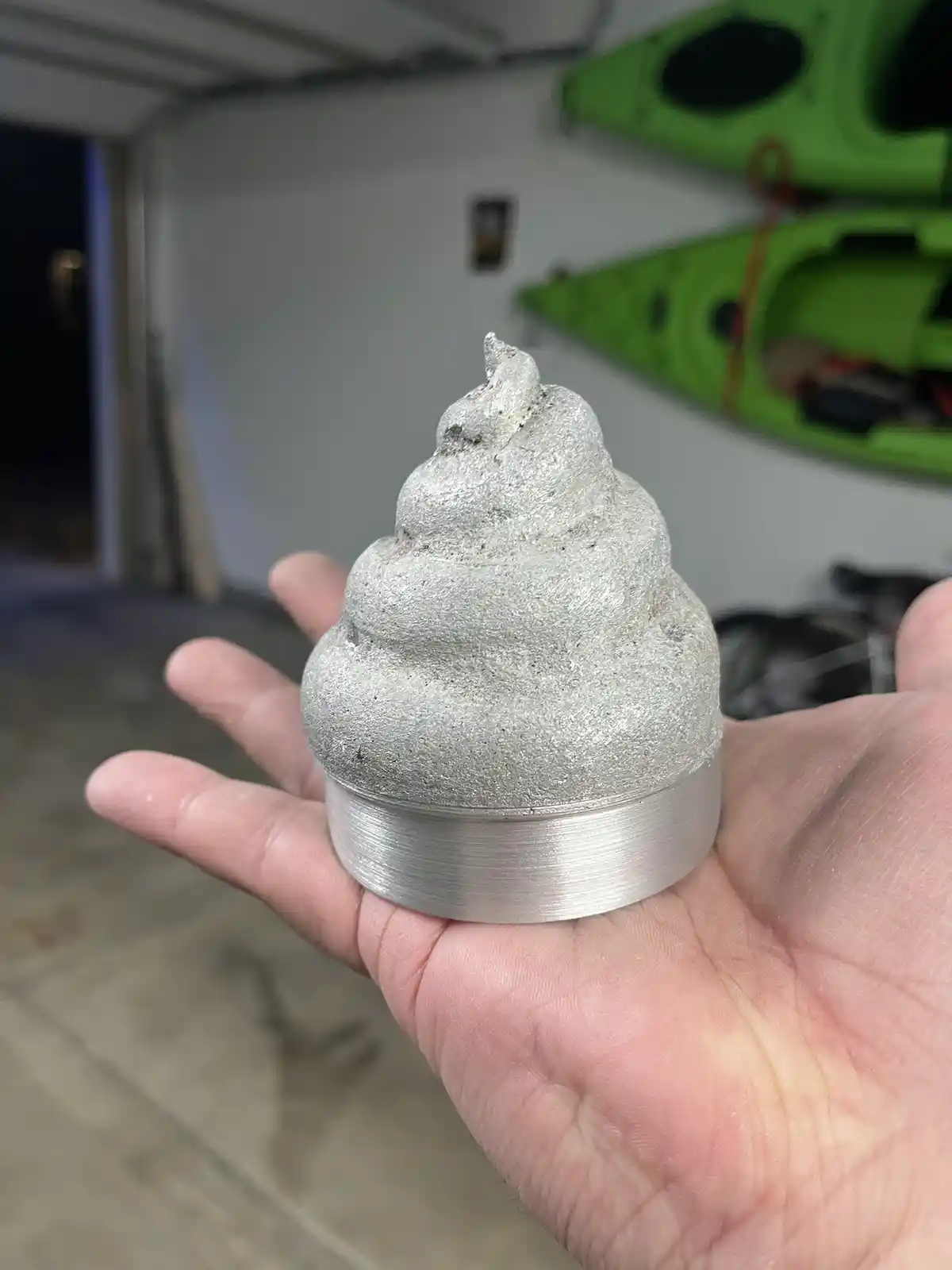 A hand holds a small, metallic object shaped like a poop emoji, with a brushed metal base and textured, silver-colored top…