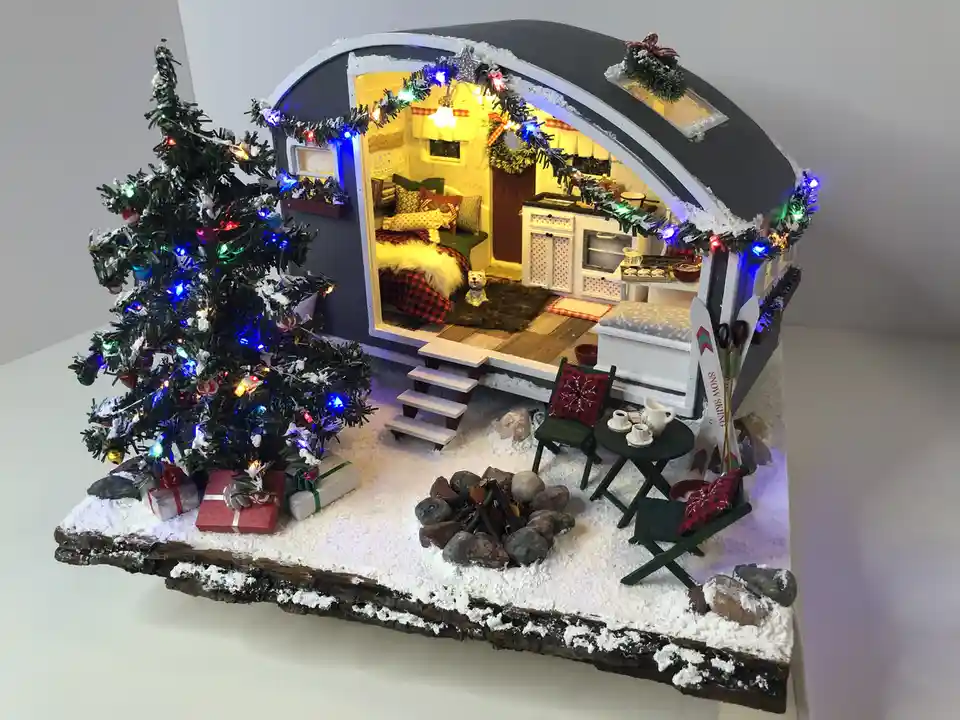 A detailed miniature diorama of a cozy, snow-covered camper decorated for Christmas, featuring a lit tree, presents, a fire…