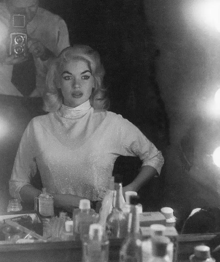 A black-and-white photo shows a woman with blonde hair and a sparkly top standing behind a cluttered table with bottles and…