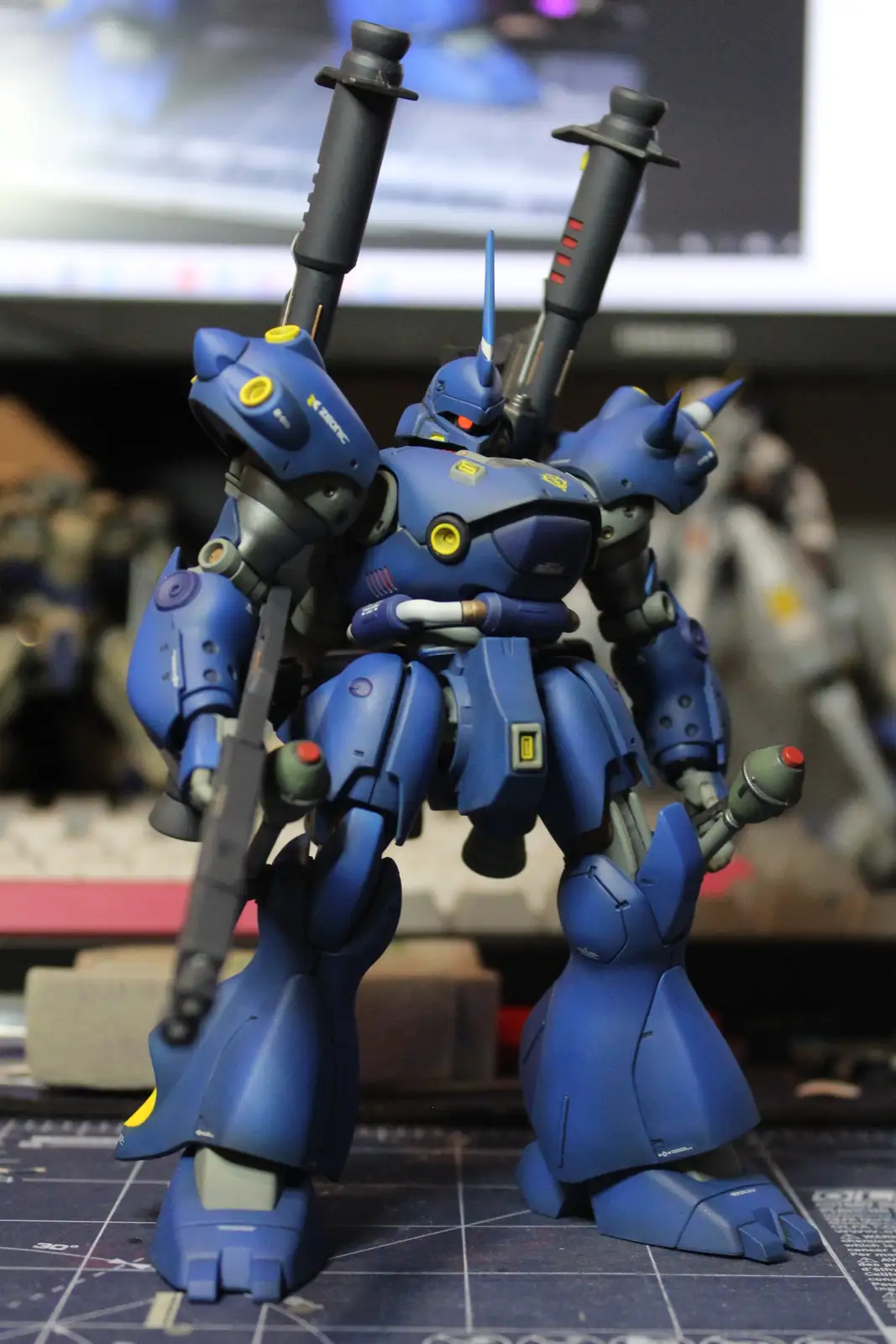 A blue, heavily armed model of a mobile suit stands on a grid-patterned mat, holding two large black weapons over its…