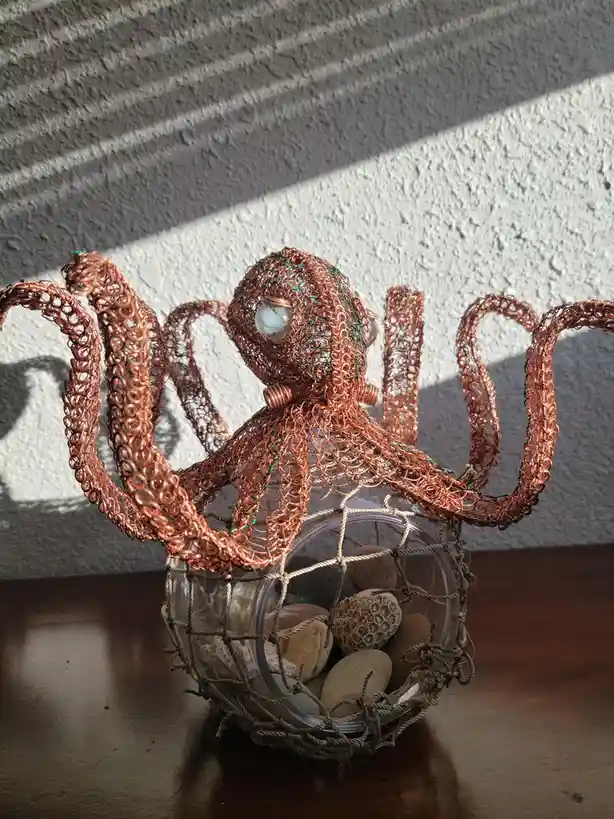 A copper wire sculpture of an octopus with large glass eyes sits inside a clear glass jar wrapped in a rope net, containing…