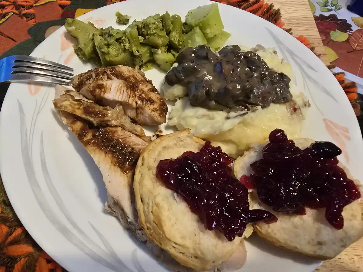 A plate of food including sliced turkey, mashed potatoes with gravy, green beans, and two biscuits topped with cranberry…
