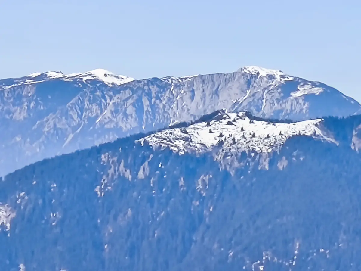 A scenic view of snow-capped mountains under a clear blue sky, with a forested ridge in the foreground. The overlaid text…