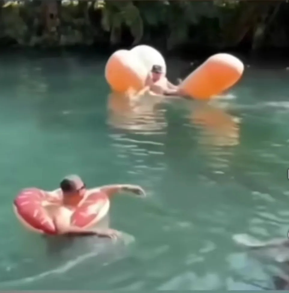 Two men are floating in greenish water, one in the foreground wearing a pink donut-shaped inflatable ring, the other further…