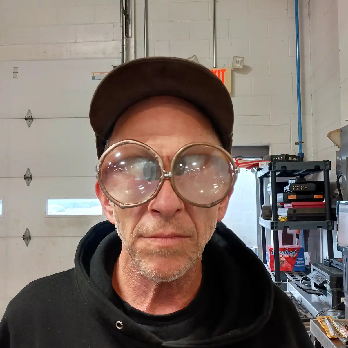 A middle-aged man with a goatee and brown cap wears absurdly oversized, round, clear-lensed glasses in a garage setting with…