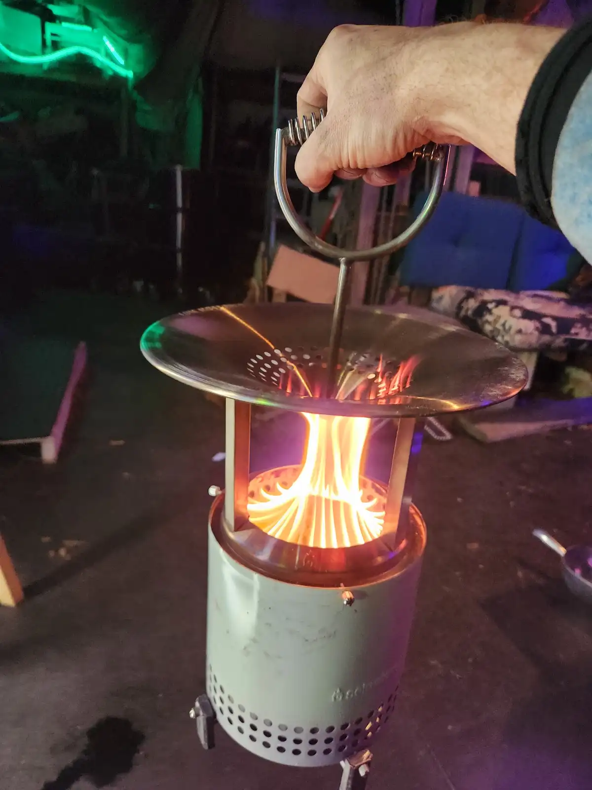 A hand holds the handle of a lit, cylindrical portable camping stove with a bright orange flame visible inside. The stove…