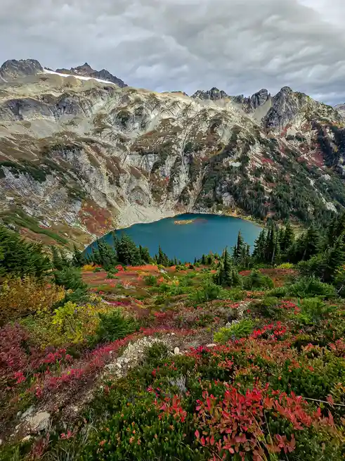 A scenic mountain landscape with a deep blue alpine lake nestled among rugged peaks under a cloudy sky. The foreground is…
