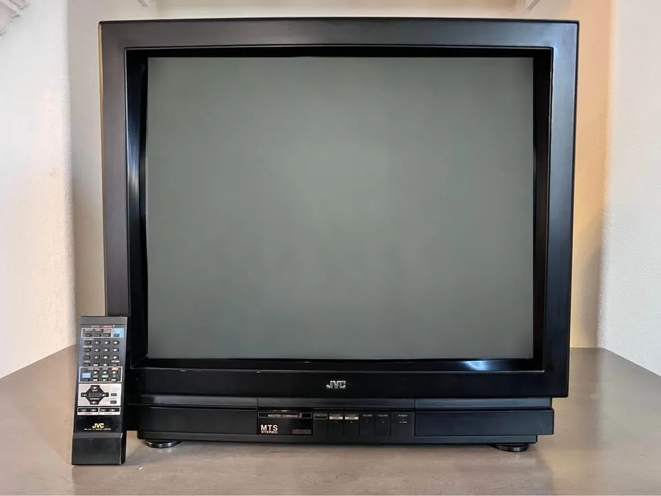 A black JVC CRT television with a blank gray screen sits on a wooden surface, accompanied by a matching JVC remote control…