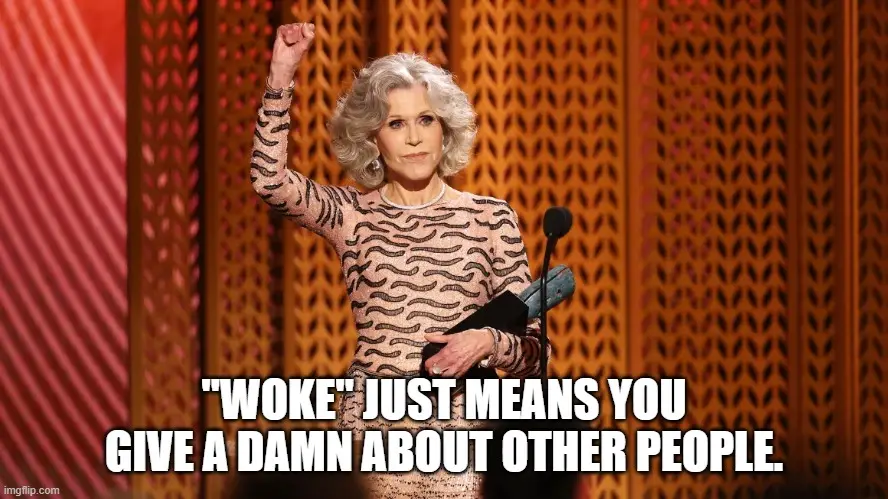 A woman with gray hair, wearing a patterned dress, stands on a stage with a microphone, raising her right fist in a…