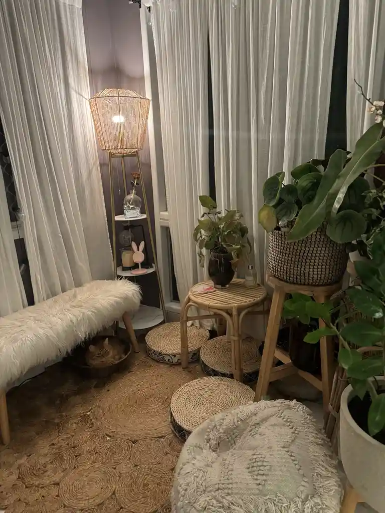 A cozy, well-lit room with white curtains, a woven rug, and multiple potted plants. A fluffy white bench sits beside a lit…