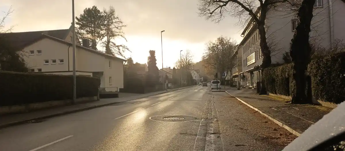 A photo of a quiet, wet street at sunrise or sunset with buildings, bare trees, and a manhole cover visible. The overlaid…