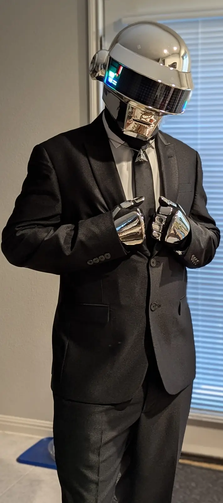A person wearing a full Daft Punk helmet with glowing visor, a black suit, white shirt, black tie, and shiny chrome gloves…
