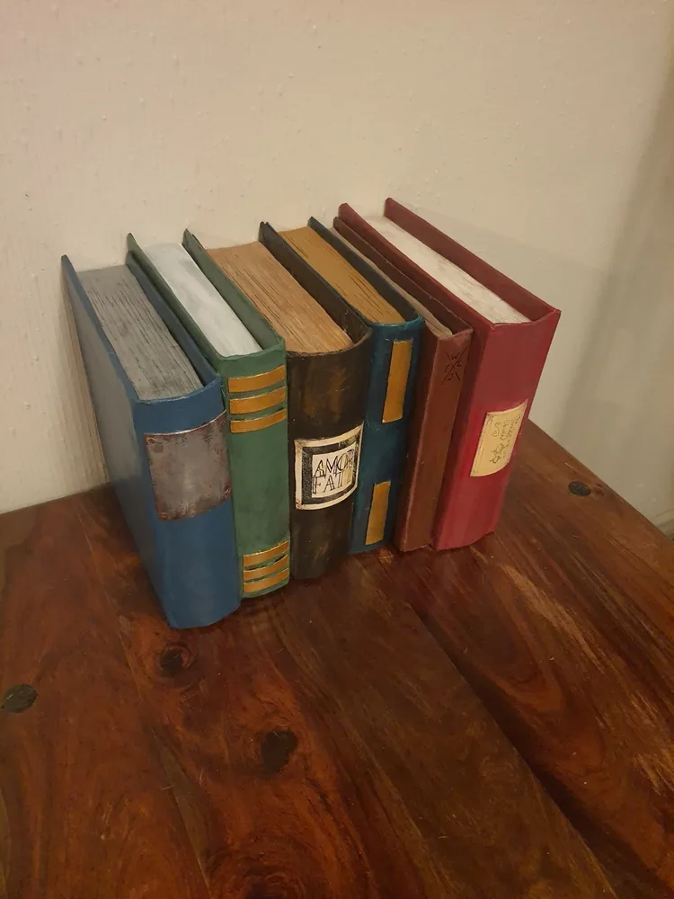 Five vintage-style books with worn spines and varied colors stand upright on a wooden surface against a textured wall. One…