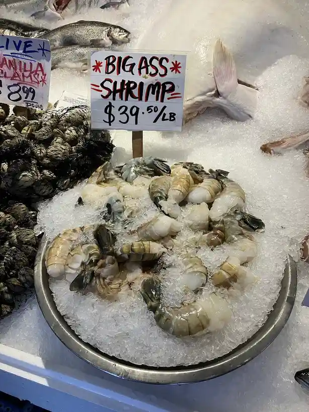A metal bowl filled with large, raw shrimp sits on crushed ice at a seafood market, with a handwritten sign above reading…