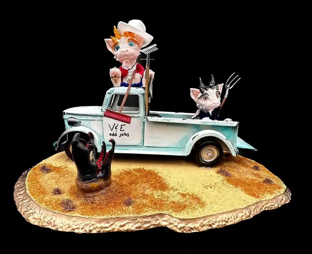 A figurine of two horned, cat-like creatures in a vintage turquoise pickup truck labeled 'V&E odd jobs'. The driver, wearing…