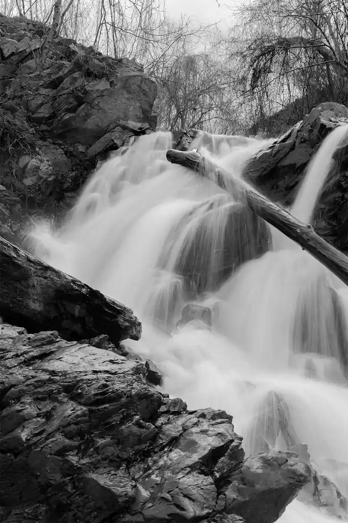 A black-and-white photograph of a waterfall with smooth, blurred water cascading over rocks, featuring a fallen log across…