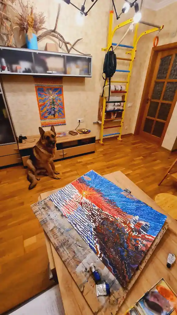 A German Shepherd dog sits patiently on a wooden floor in a room that doubles as an art studio and home gym, with a large…