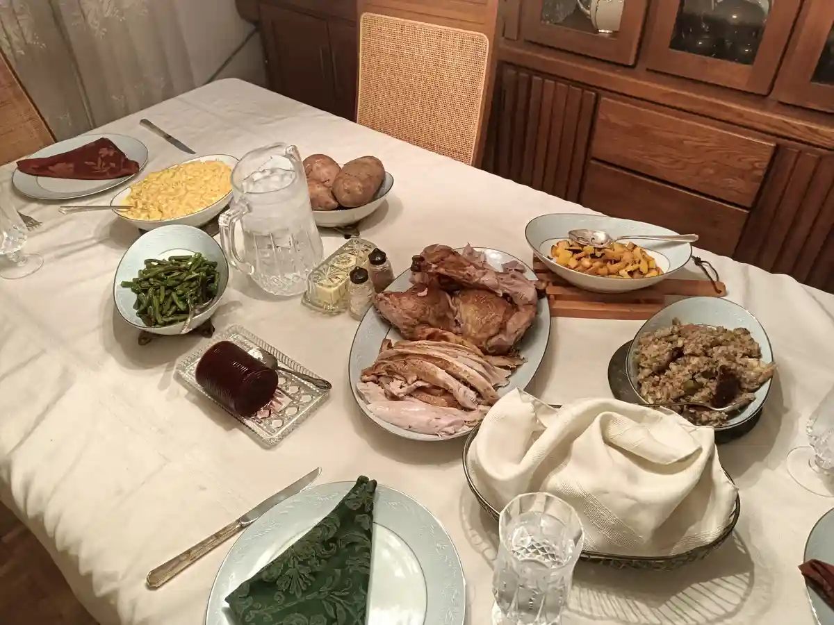 A dining table is set with a large roasted turkey, macaroni and cheese, green beans, sweet potatoes, stuffing, and cranberry…