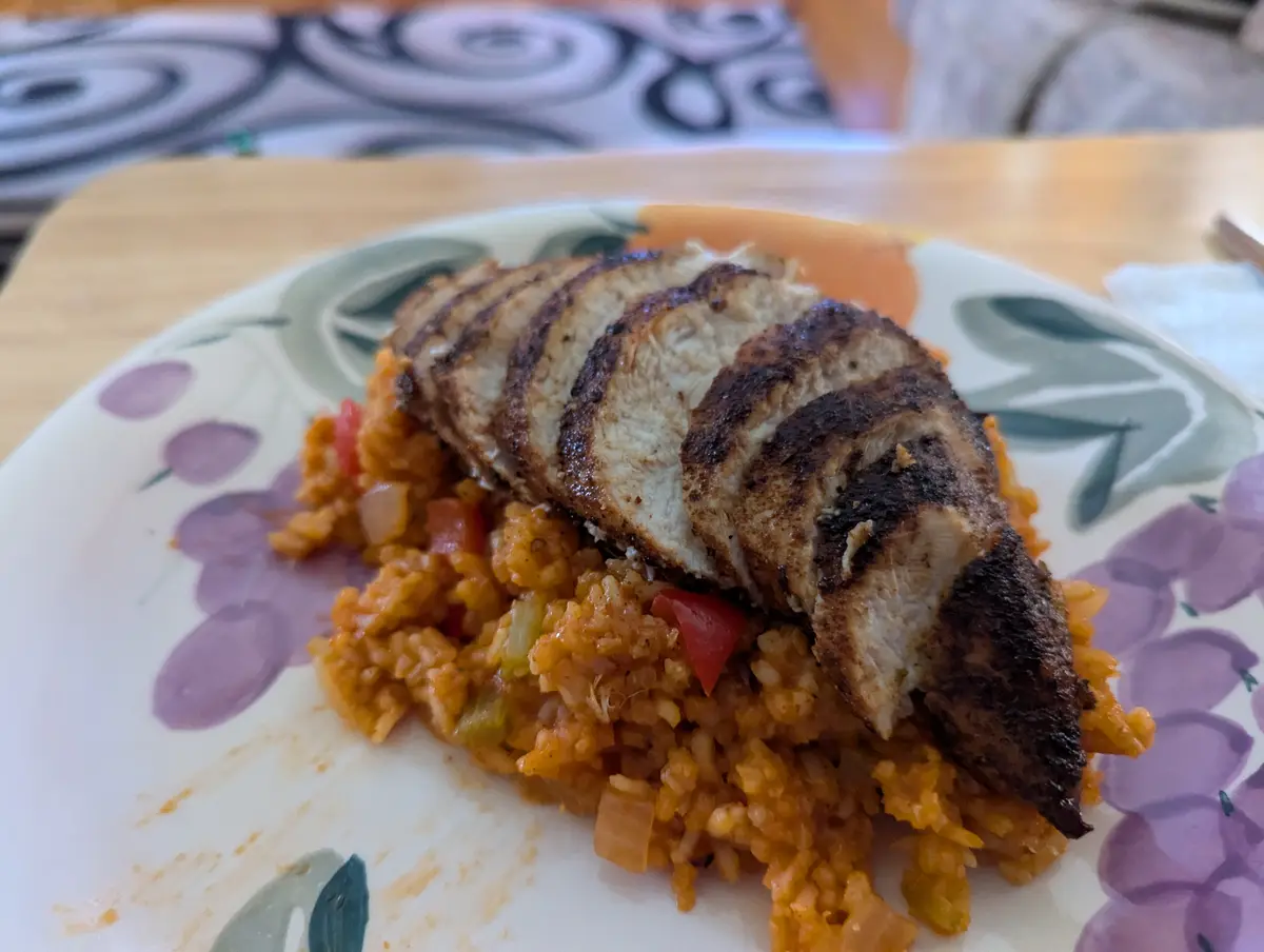A close-up photo of a plate of rice with sliced grilled chicken on top, the chicken has distinct grill marks and appears…
