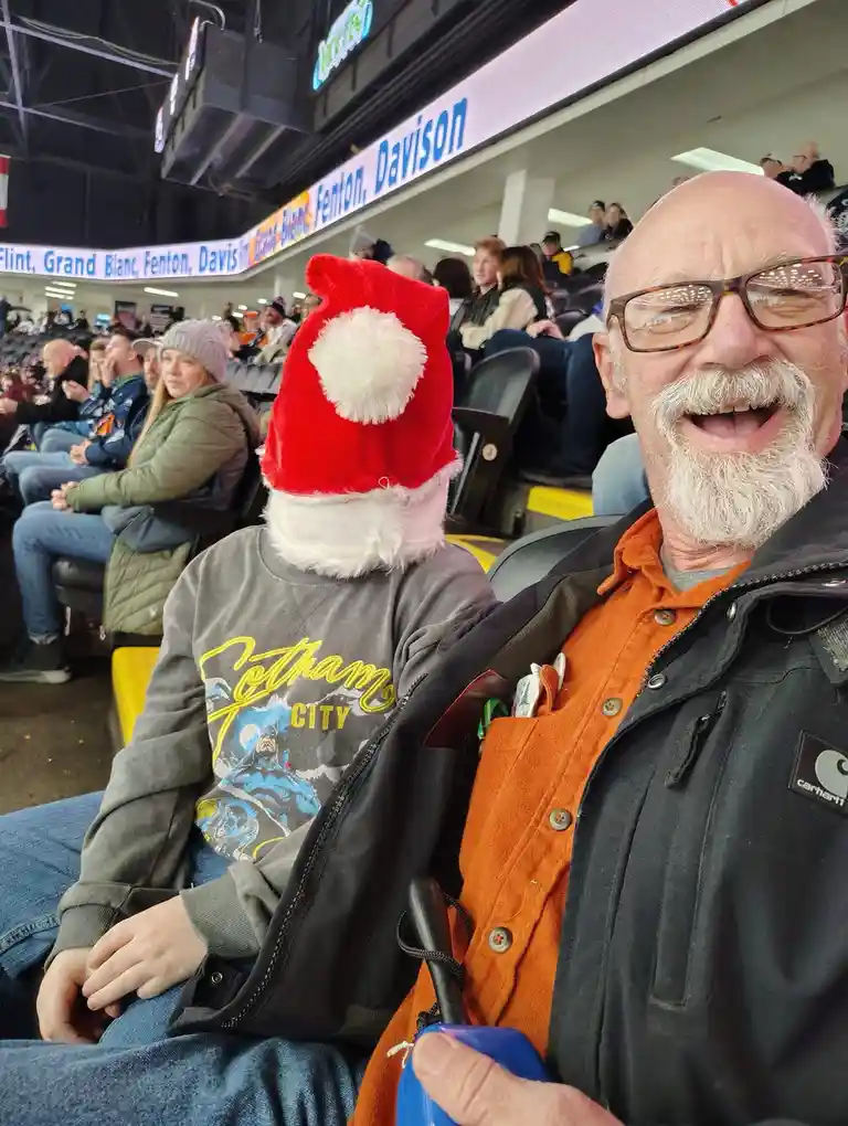 A man with glasses and a white beard laughs broadly while sitting in a stadium, next to another person wearing a large red…