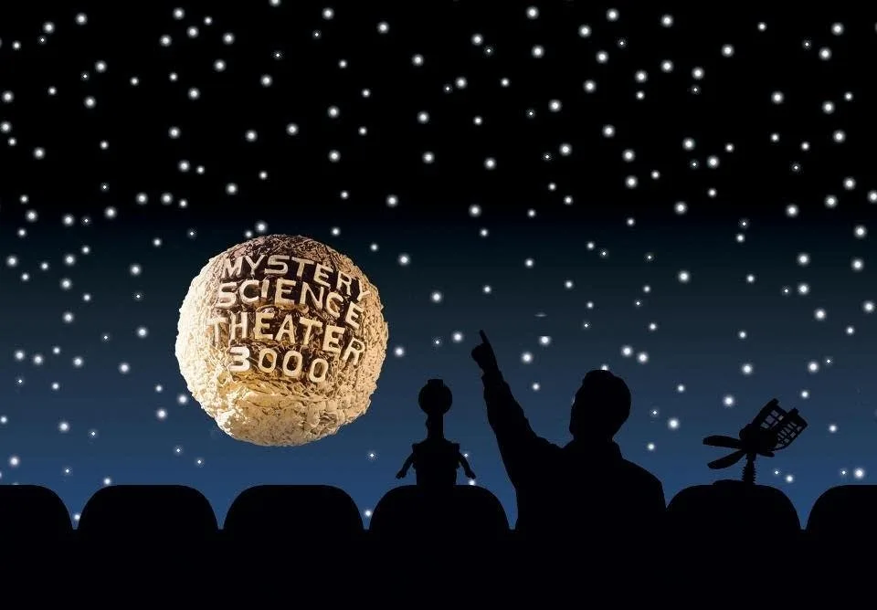 A silhouette of a person and two robots sitting in theater seats, pointing upwards at a large, textured moon against a…